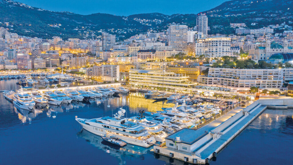 Luxury yachts moored at a brightly lit marina in Monaco, surrounded by city buildings at dusk. Yacht club